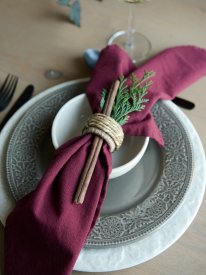 Närbild på en stämningsfull dukning i lantlig stil med en djupt vinröd tygservett Vide från Fondaco dekorerad med en kanelstång 