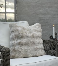 Fluffigt kuddfodral Matteo Sand 48x48 cm från Fondaco i fuskpäls placerat i en rottingfåtölj på en mysig uteplats med levande lj