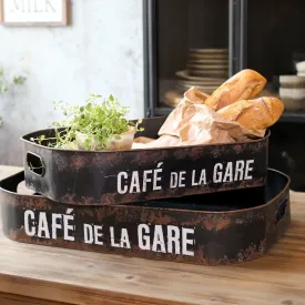 Två staplade antiksvarta metallbrickor Café de la Gare med fransk text, stylade med färskt bröd och en grön örtkruka på ett rust