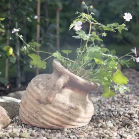 Liggande antik terrakottaurna Talia från Wikholm Form placerad på en grusbädd med rosa blommor som växer ut ur öppningen.