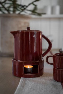 A red mulled wine pitcher with a warmer and a lit candle inside, on a rustic-style Christmas table.