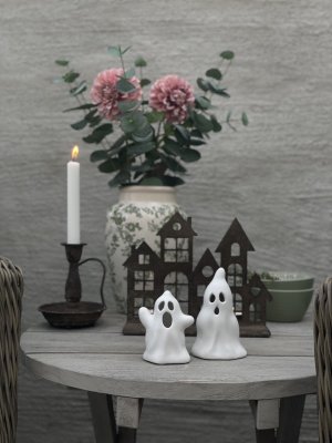 Two small white ceramic ghosts serving as candle holders, beautifully styled on a round wooden table in front of a rusty decorat