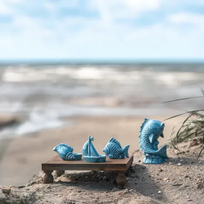 Den blå keramikfisken ståendes i den ljusa sanden på en strand tillsammans med andra blå maritima detaljer i keramik.