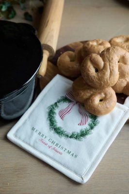 Julig miljöbild med en offwhite grytlapp Ribbon med julkransmotiv, placerad på en köksbänk bredvid en hög med nybakade lussekatt