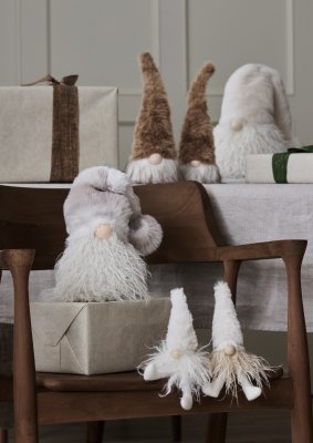Mjuk tomte Sten i beigemelerad fejkpels sitter på ett julpaket på en trästol omgiven av andra mjuka tomtar och julklappar från W
