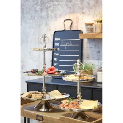 Lyxigt etagefat i silverfärgad aluminium fyllt med tapas, ostar och charkuterier på ett serveringsbord i trä.